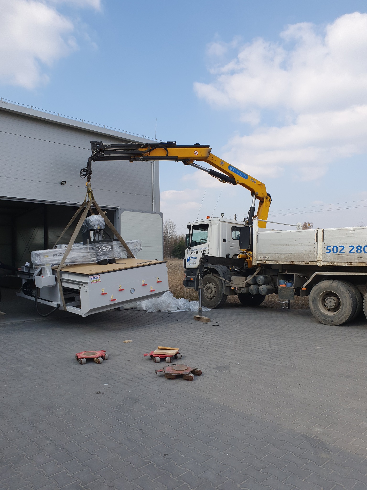 Żółty dźwig samochodowy podnosi dużą, białą maszynę CNC z hali magazynowej na zewnątrz, na kostkę brukową, z użyciem pasów transportowych.