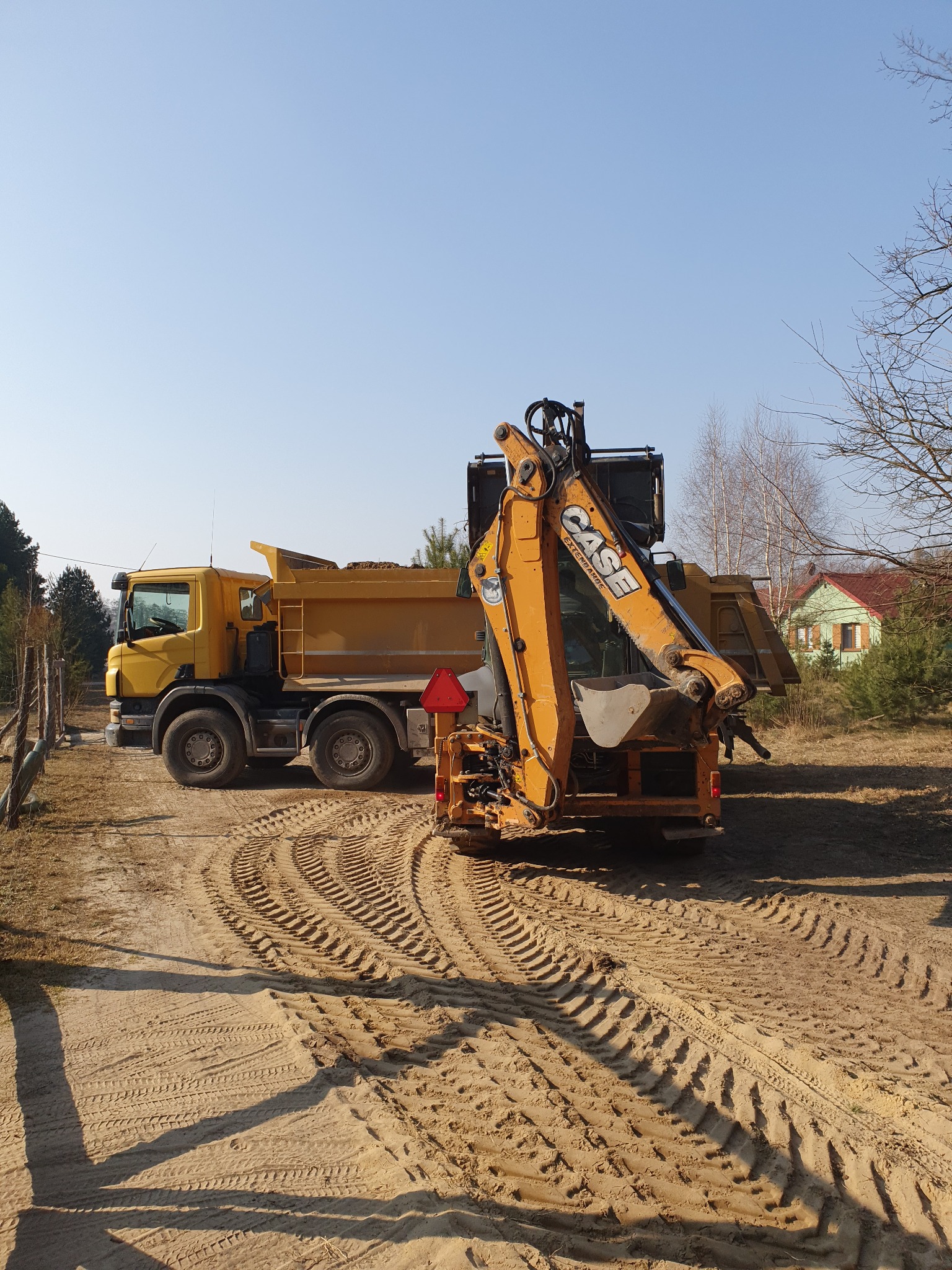 Żółta wywrotka załadowana piaskiem i pomarańczowa koparko-ładowarka CASE na piaszczystym terenie, ślady opon widoczne na ziemi, w tle budynek z zielonymi ścianami i czerwonym dachem.