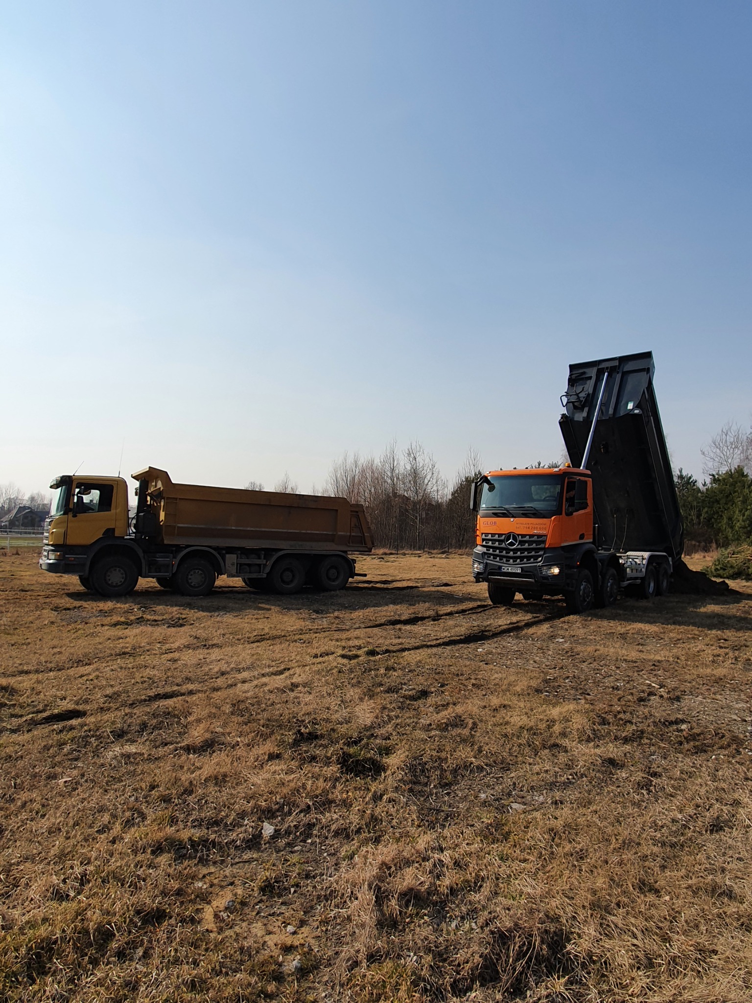 Dwa samochody ciężarowe na polu: żółty wywrotka i pomarańczowy wywrotka z uniesioną skrzynią ładunkową, wysypujący materiał na ziemię.