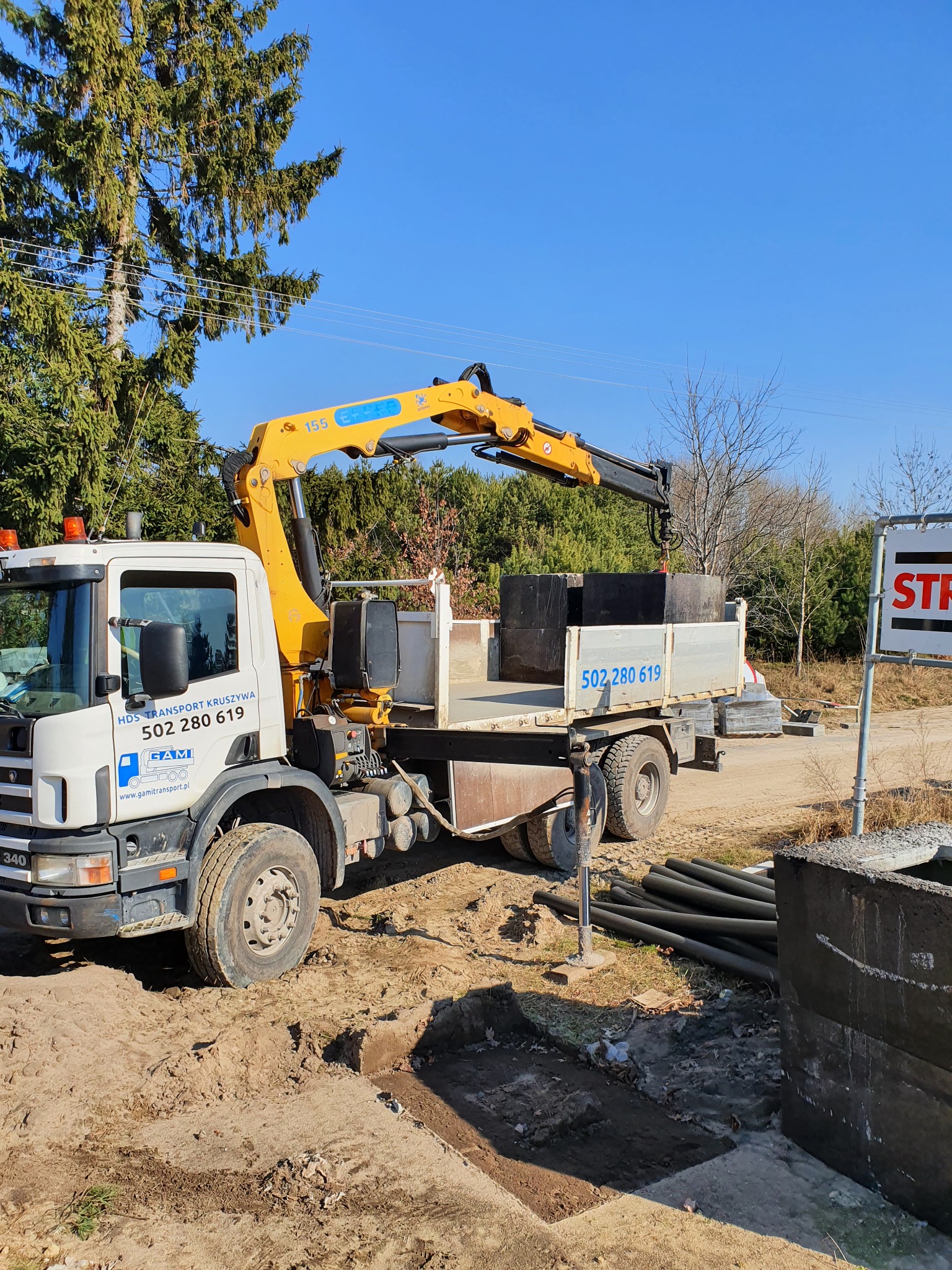 Ciężarówka z dźwigiem transportuje betonowe elementy na placu budowy, rury kanalizacyjne obok wykopu, w tle drzewa i błękitne niebo.