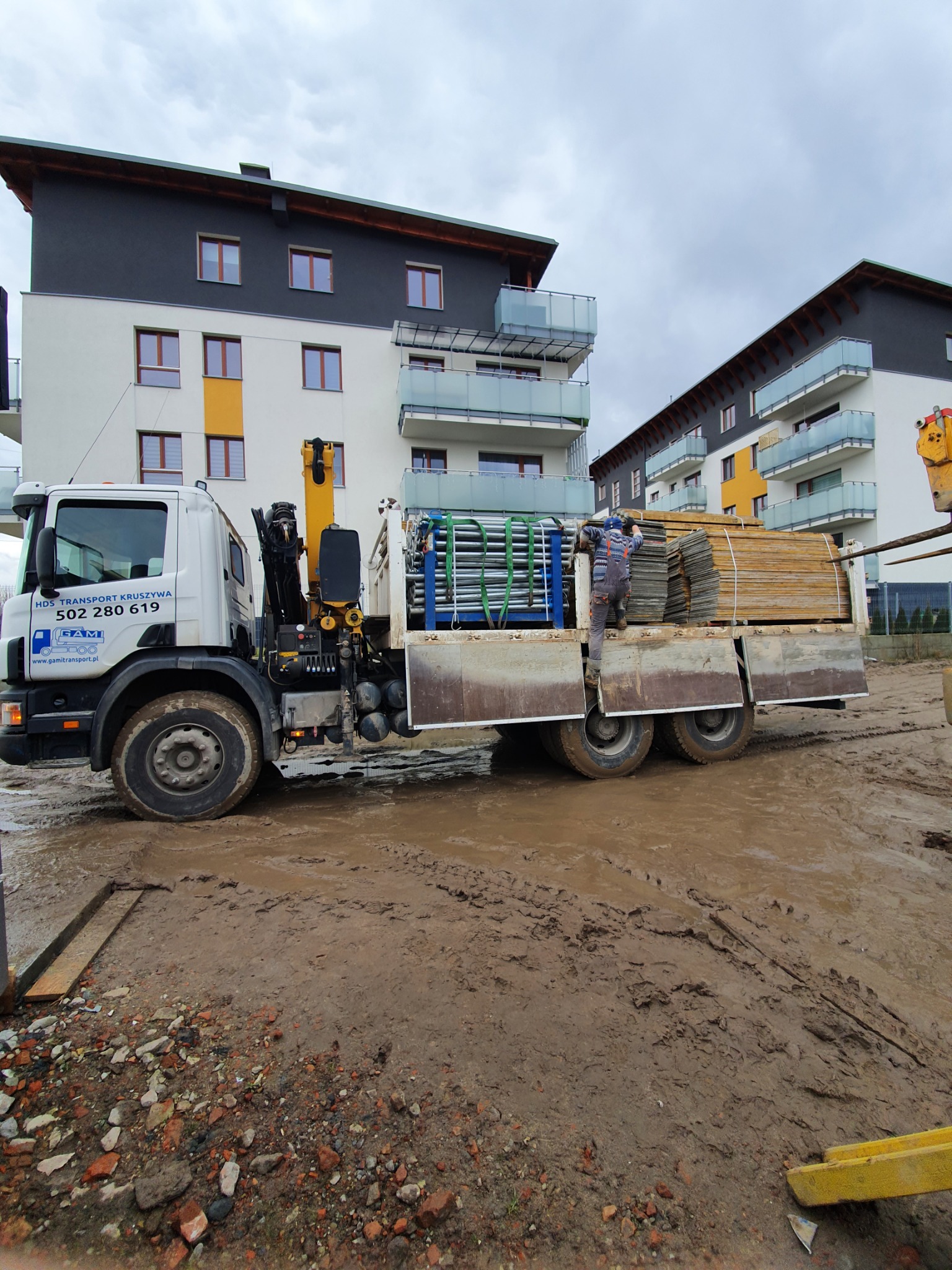 Ciężarówka HDS firmy transportowej HDS Transport Kruszywa z załadunkiem płyt betonowych i rusztowań na błotnistym placu budowy, w tle budynki mieszkalne.