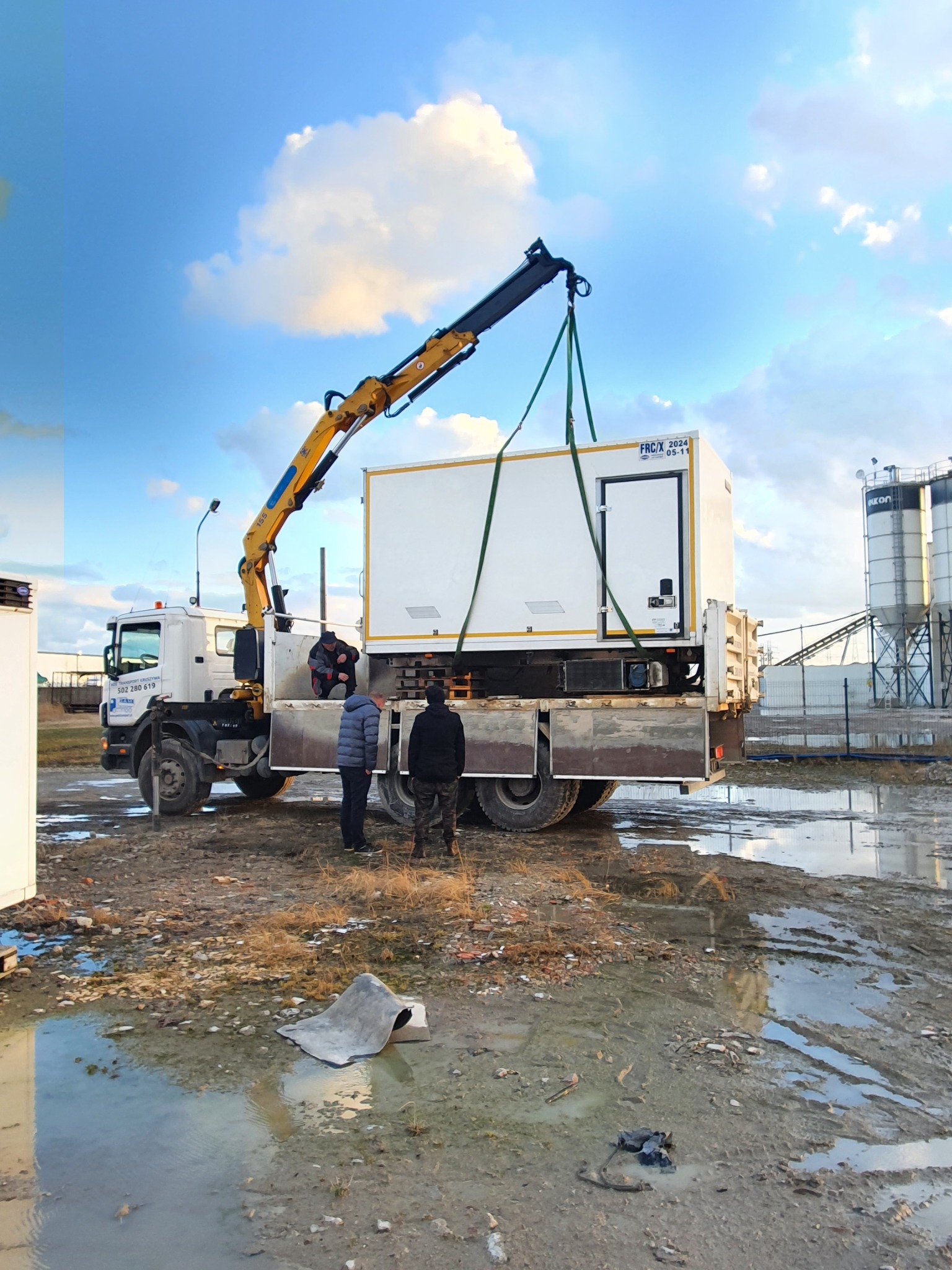 Żuraw samochodowy podnosi biały kontener na platformę ciężarówki w błotnistym terenie, w tle widoczne silosy i pochmurne niebo.