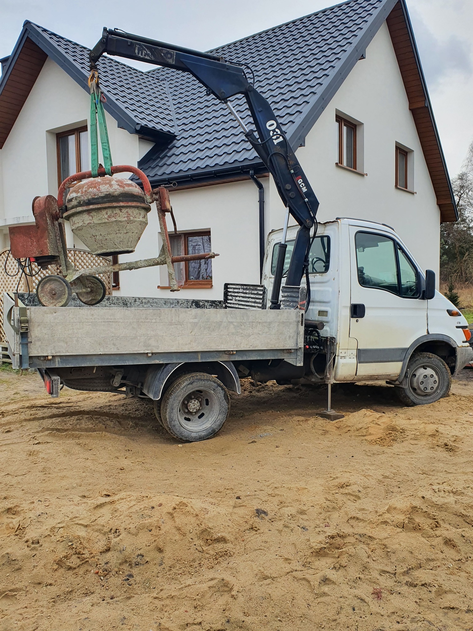 Ciężarówka z dźwigiem podnosi brudną betoniarkę na tle białego domu z ciemną dachówką.