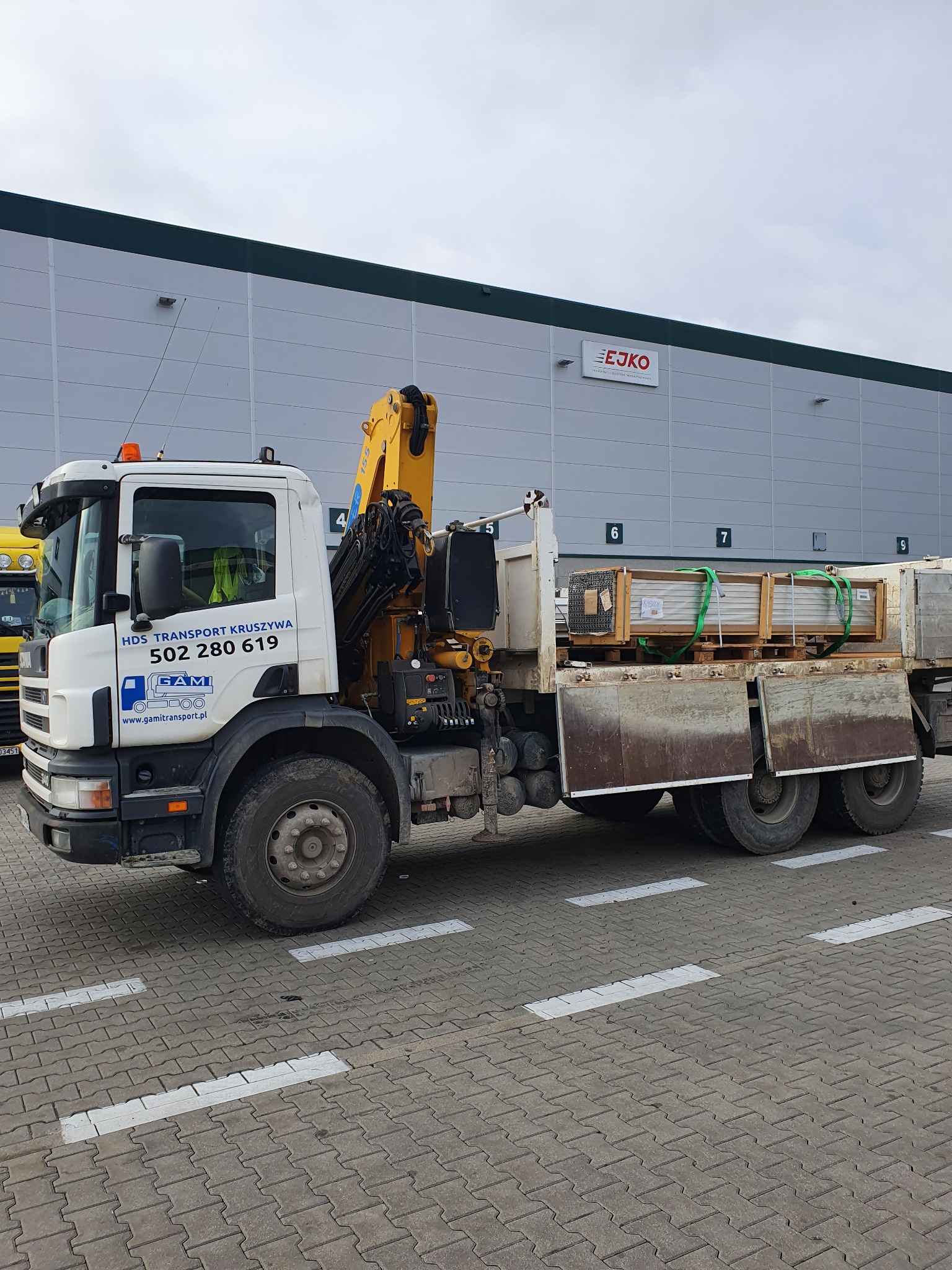 Ciężarówka z dźwigiem HDS firmy HDS Transport Kruszywa z załadowanymi prefabrykatami betonowymi na paletach, zabezpieczonymi zielonymi pasami transportowymi, na tle hali magazynowej z logo firmy EJKO.