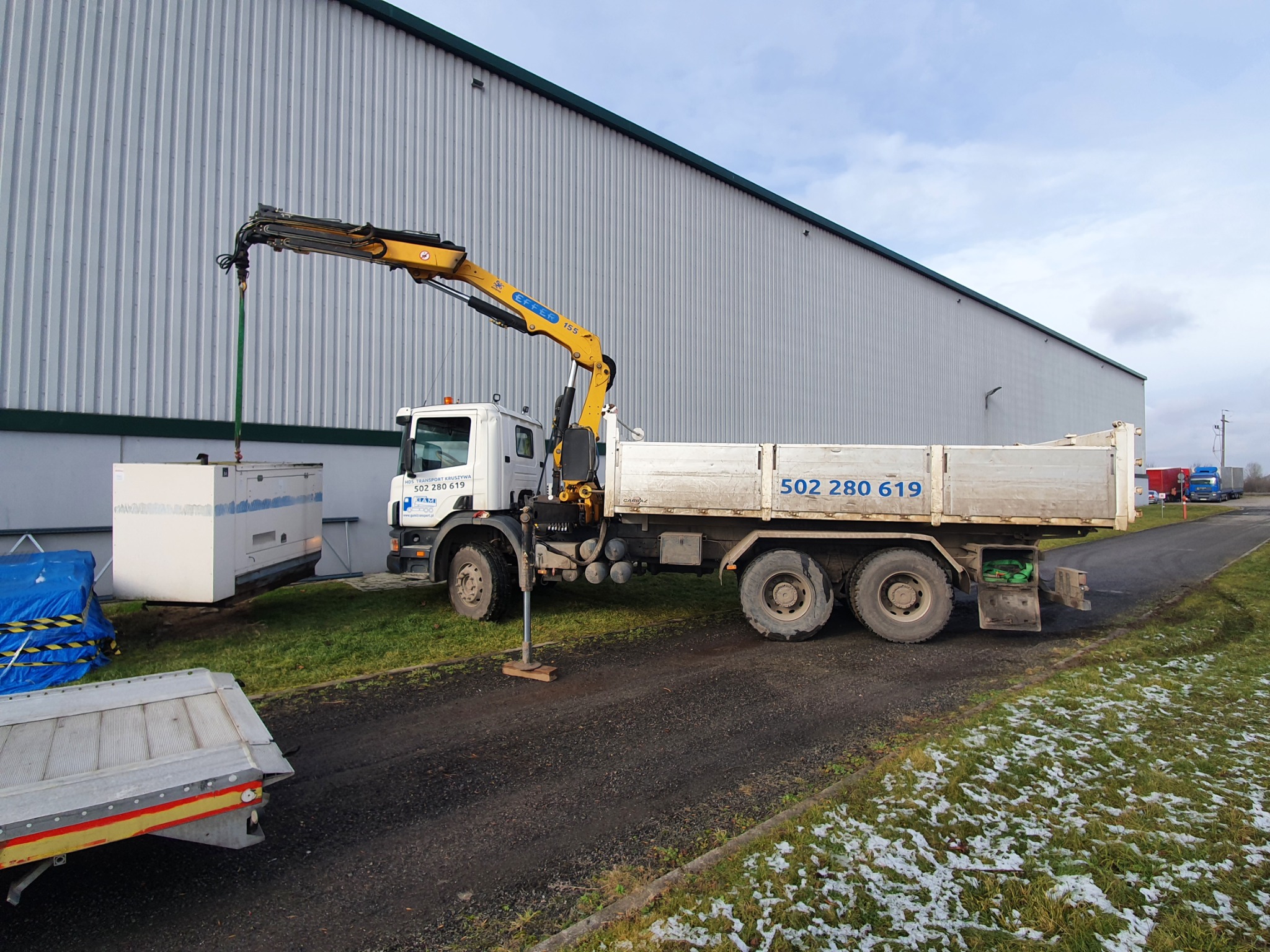 Ciężarówka z dźwigiem przenosi biały agregat prądotwórczy na tle hali magazynowej, widoczny numer telefonu na burcie