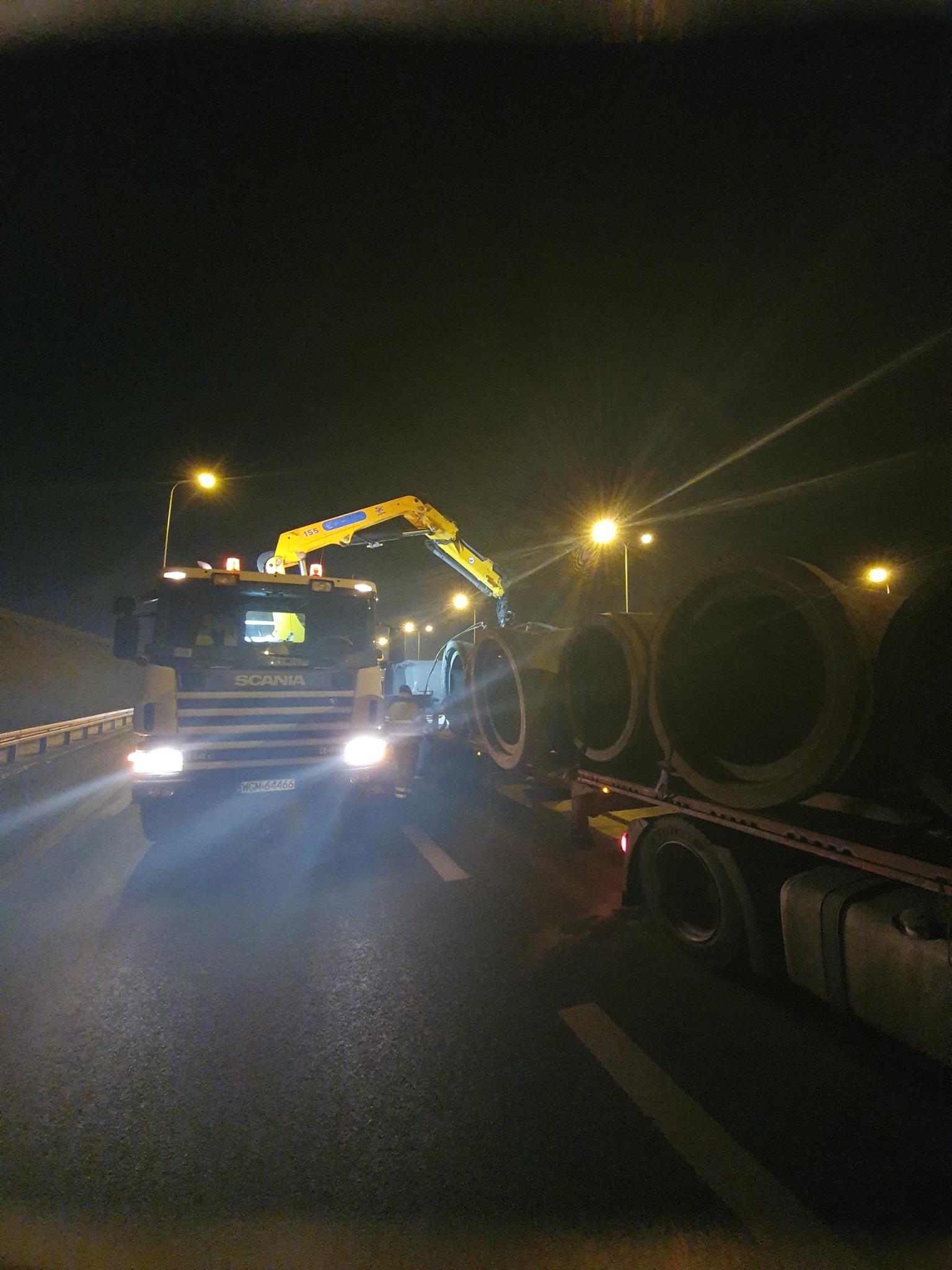 Ciężarówka SCANIA z żółtym dźwigiem przewozi betonowe kręgi na platformie przy oświetlonej ulicy nocą.