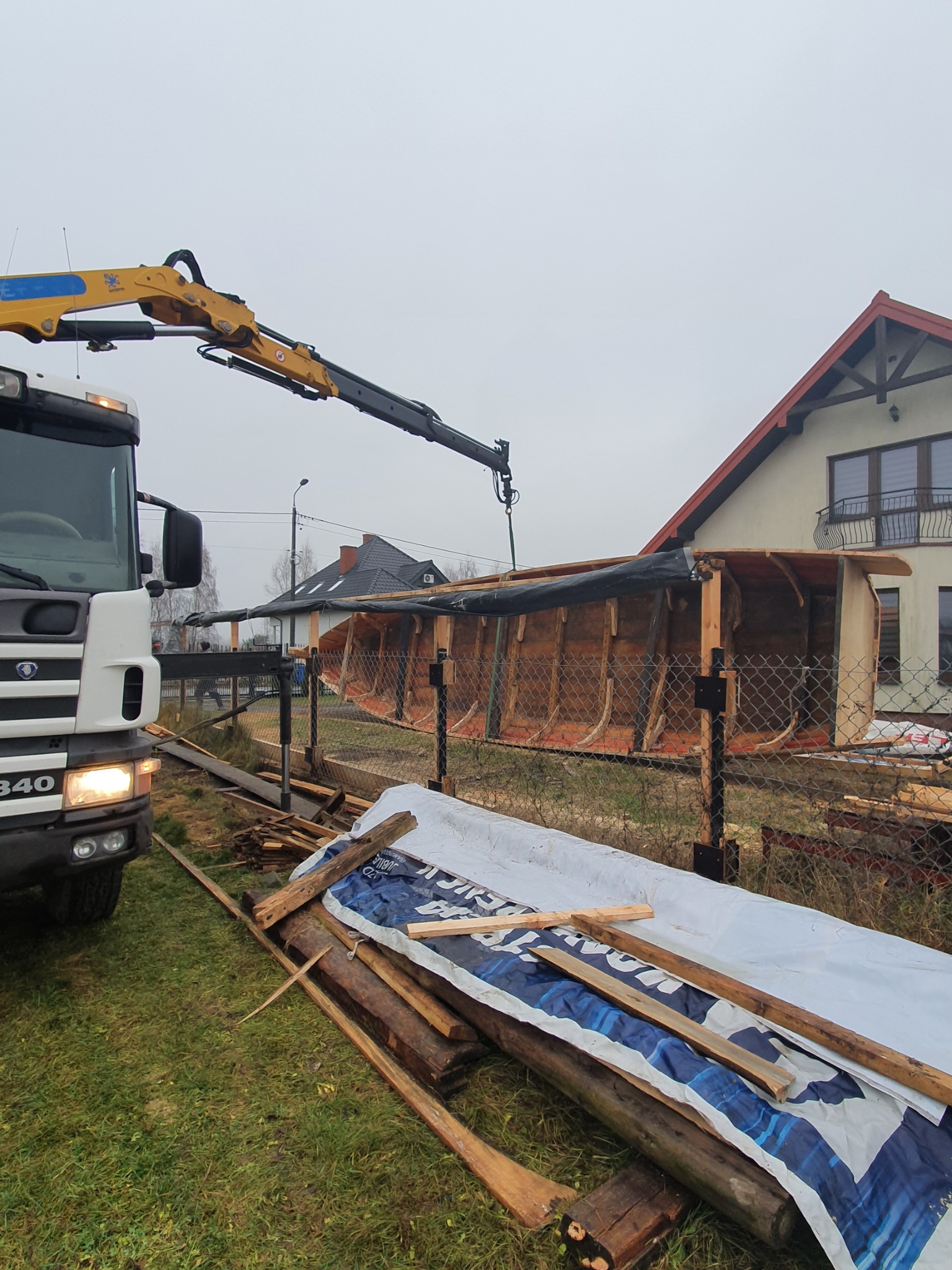 Żuraw samochodowy podnosi drewnianą konstrukcję szkieletową przy ogrodzeniu z siatki, w tle dom jednorodzinny, pochmurne niebo.