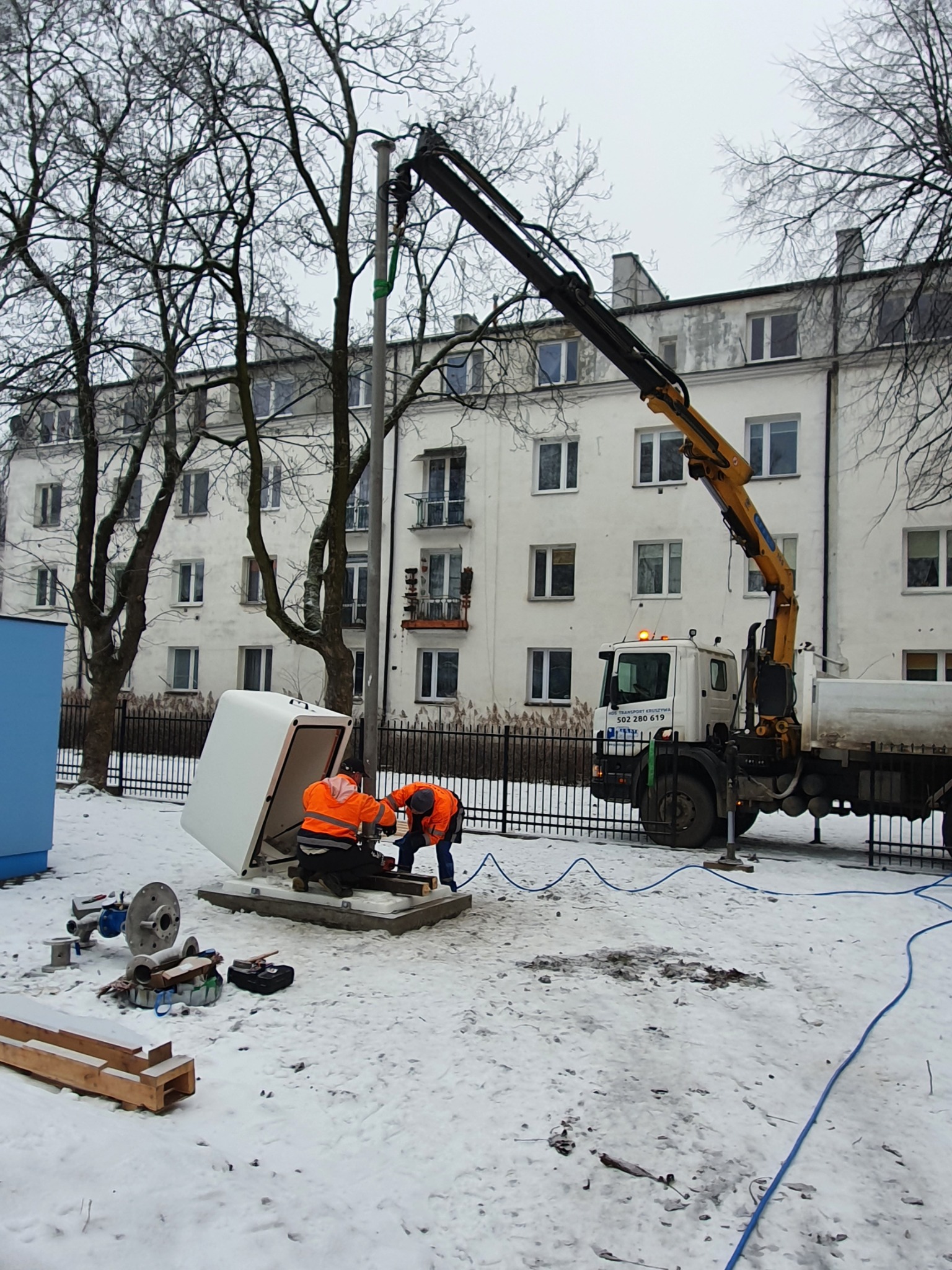 Montaż białej szafy technicznej na betonowej podstawie w zimowych warunkach; dwóch pracowników w pomarańczowych kamizelkach instaluje urządzenie przy użyciu dźwigu samochodowego na tle budynku...