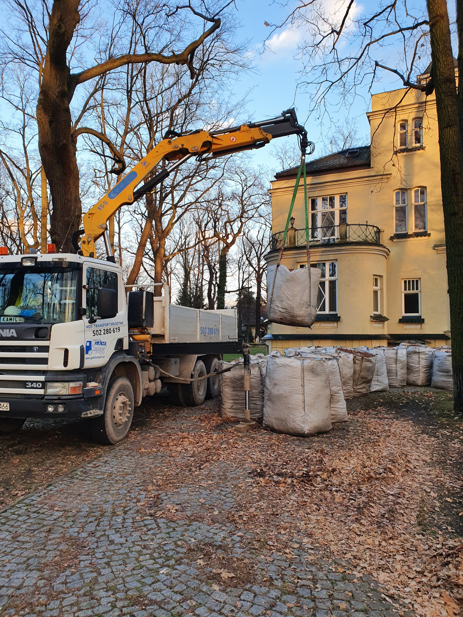 Ciężarówka z dźwigiem podnosi worki z liśćmi na tle budynku z balkonem, jesienne liście na brukowanej drodze.