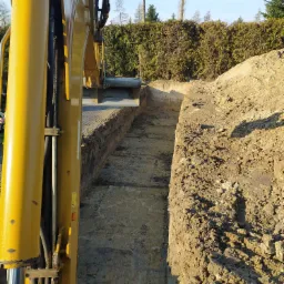 Żółta koparka z łyżką w trakcie wykonywania wykopu o równych ścianach, widoczne warstwy ziemi i roślinność w tle.