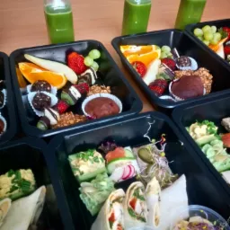 Zestaw posiłków w czarnych pojemnikach: sałatki, tortille i desery owocowe, uzupełnione butelkami zielonego soku, ułożone na drewnianym stole.