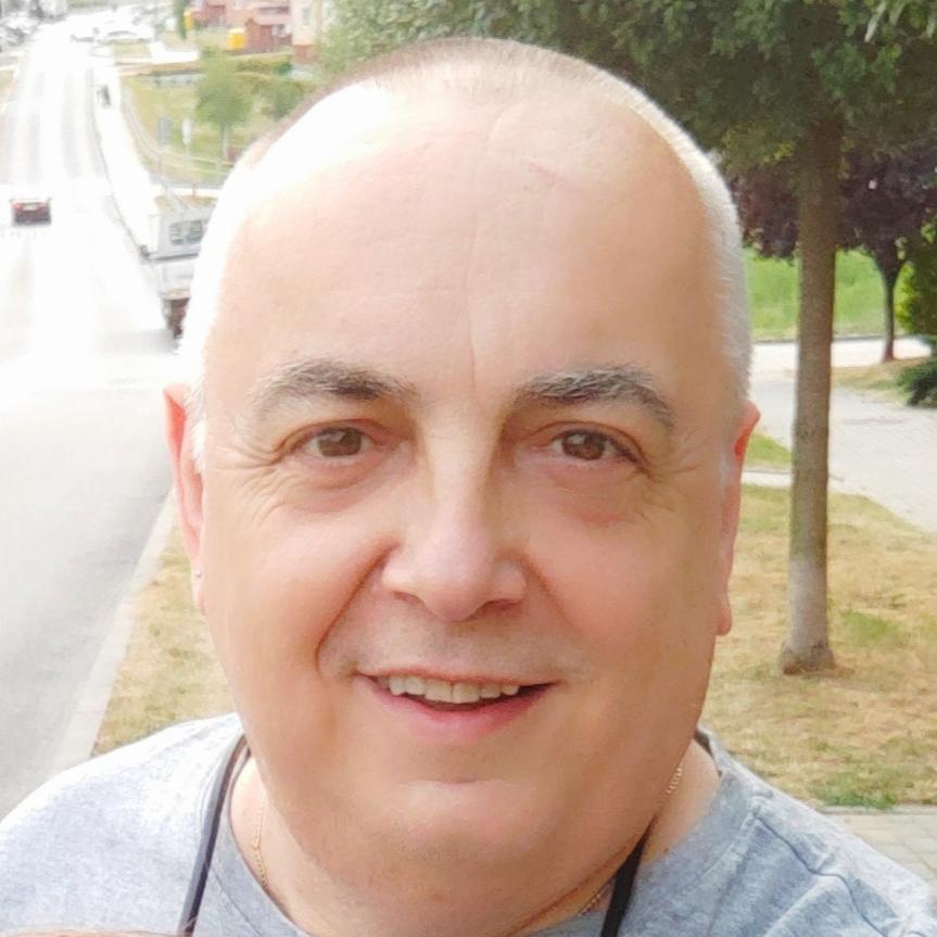 Uśmiechnięty mężczyzna z krótkimi, siwiejącymi włosami, na tle ulicy i zieleni, z widocznym fragmentem złotego łańcuszka. Portret wykonany z bliska.