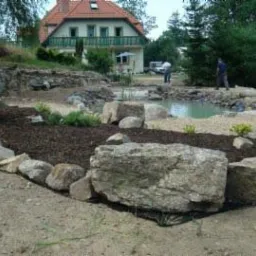 Aranżacja ogrodu z oczkiem wodnym, brzeg wyłożony kamieniami, w tle dom z balkonem i dwoma osobami pracującymi nad wykończeniem terenu.