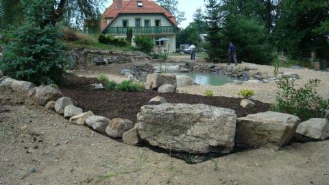 Aranżacja ogrodu z oczkiem wodnym, brzeg wyłożony kamieniami, w tle dom z balkonem i dwoma osobami pracującymi nad wykończeniem terenu.