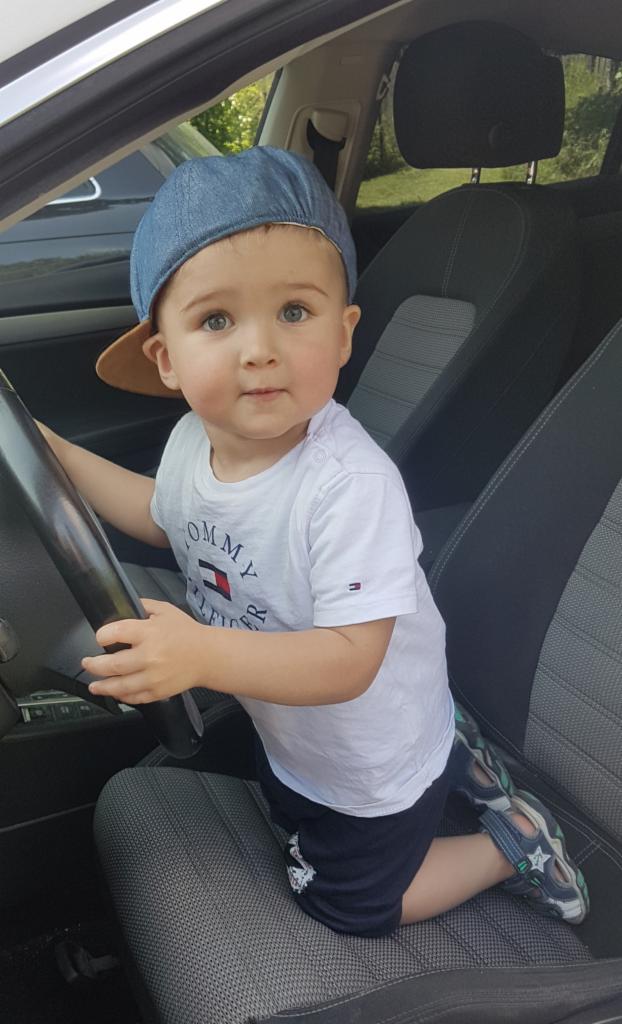 Mały chłopiec w dżinsowej czapce siedzi za kierownicą samochodu, spoglądając w obiektyw.