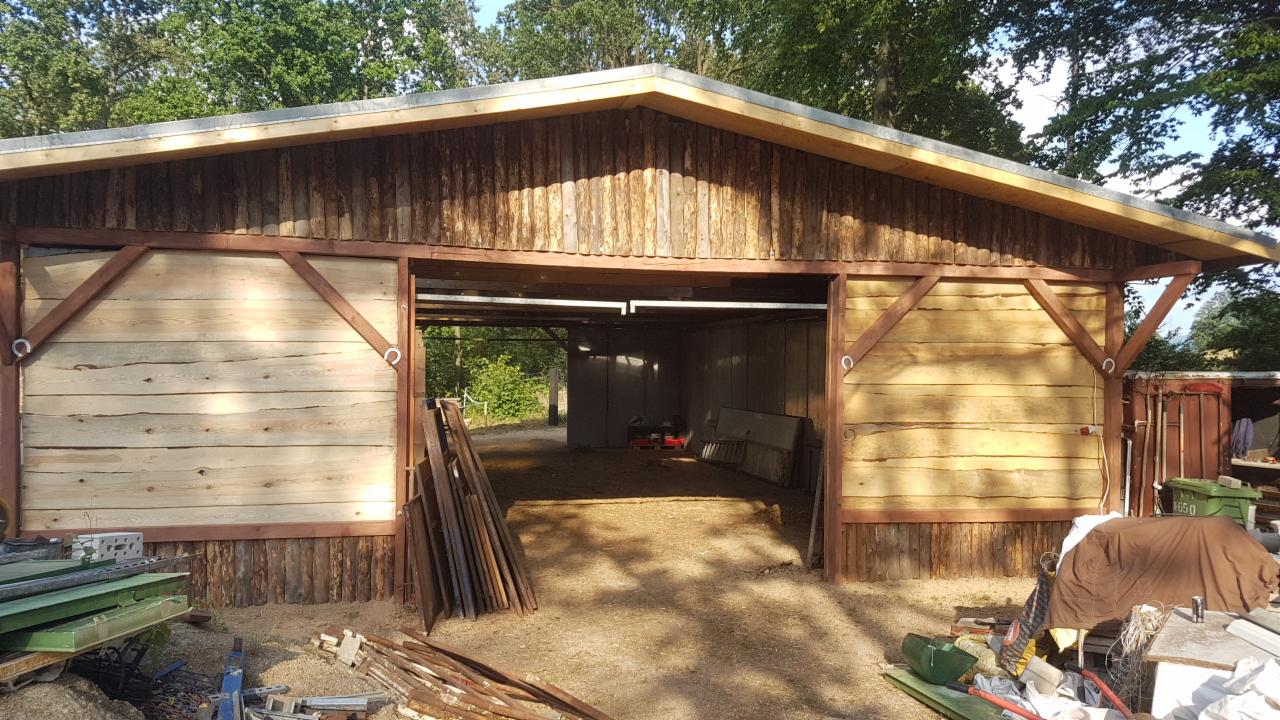 Drewniana konstrukcja stodoły z otwartymi wrotami, widoczne elementy drewniane, konstrukcja dachu oraz nieporządek wokół budowli.