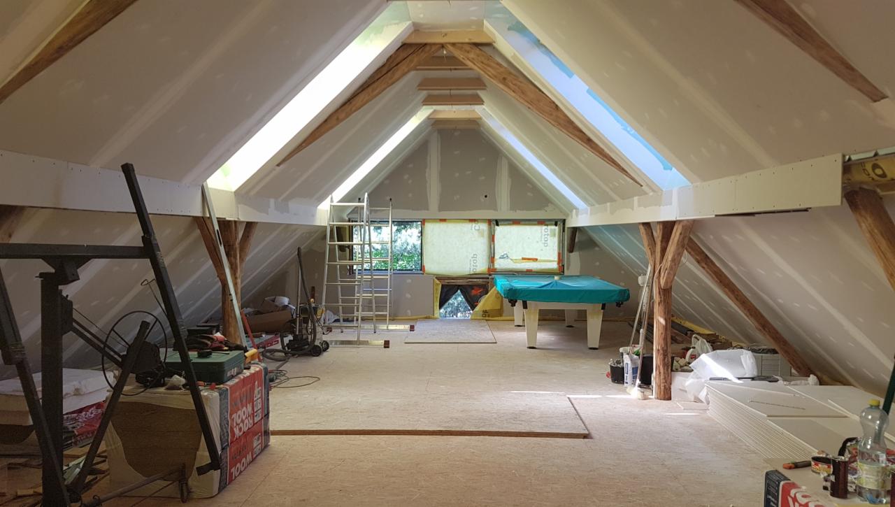 Adaptacja poddasza: widok na ocieplone skosy, częściowo wykończone płyty gipsowo-kartonowe, drewniane belki konstrukcyjne i okno dachowe, z widocznym stołem bilardowym w tle.