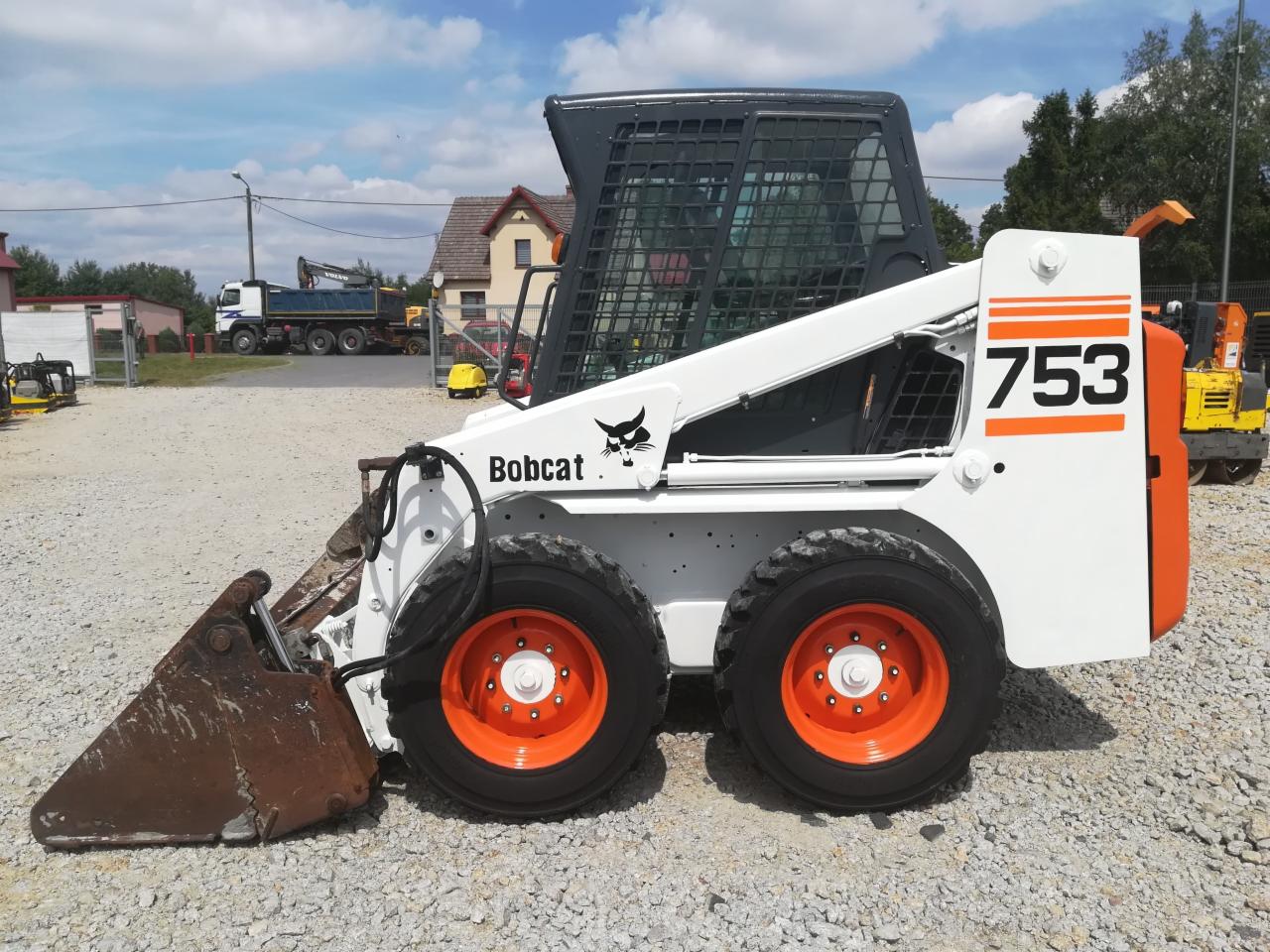 Kompaktowa ładowarka Bobcat 753 z pomarańczowymi felgami, widoczny lekko zardzewiały osprzęt roboczy, na żwirowym placu z budynkami i innymi maszynami w tle.