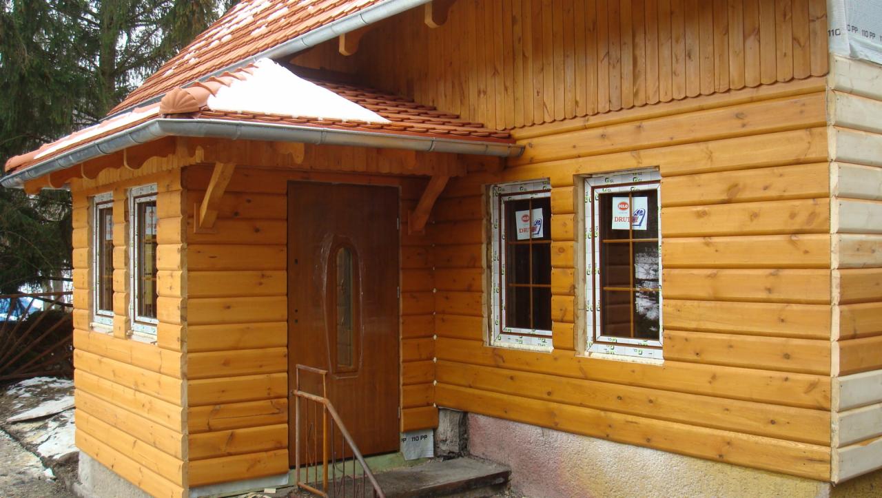 Drewniany domek z nową elewacją z desek i oknami z plastikowymi ramami, częściowo pokryty śniegiem.