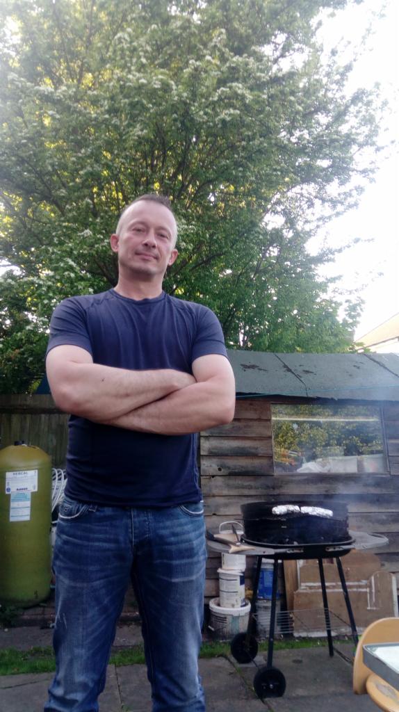 Mężczyzna w dżinsach i ciemnej koszulce stoi na tle grilla, szopy i drzewa. Na grillu leżą zawinięte w folię produkty.