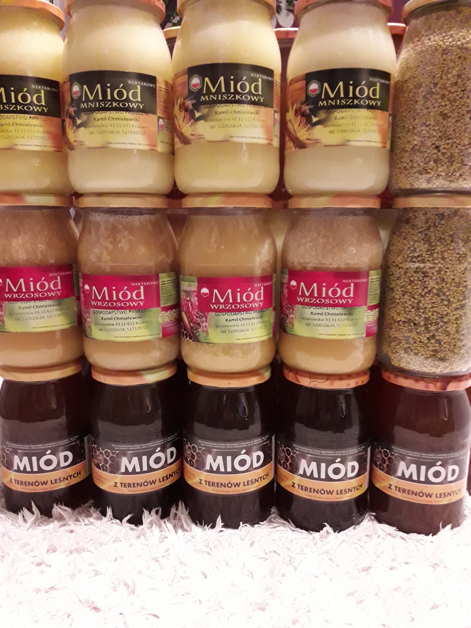 Piramida ułożona z słoików z różnymi rodzajami miodu: leśnym, wrzosowym i mniszkowym na białym, puszystym podłożu.