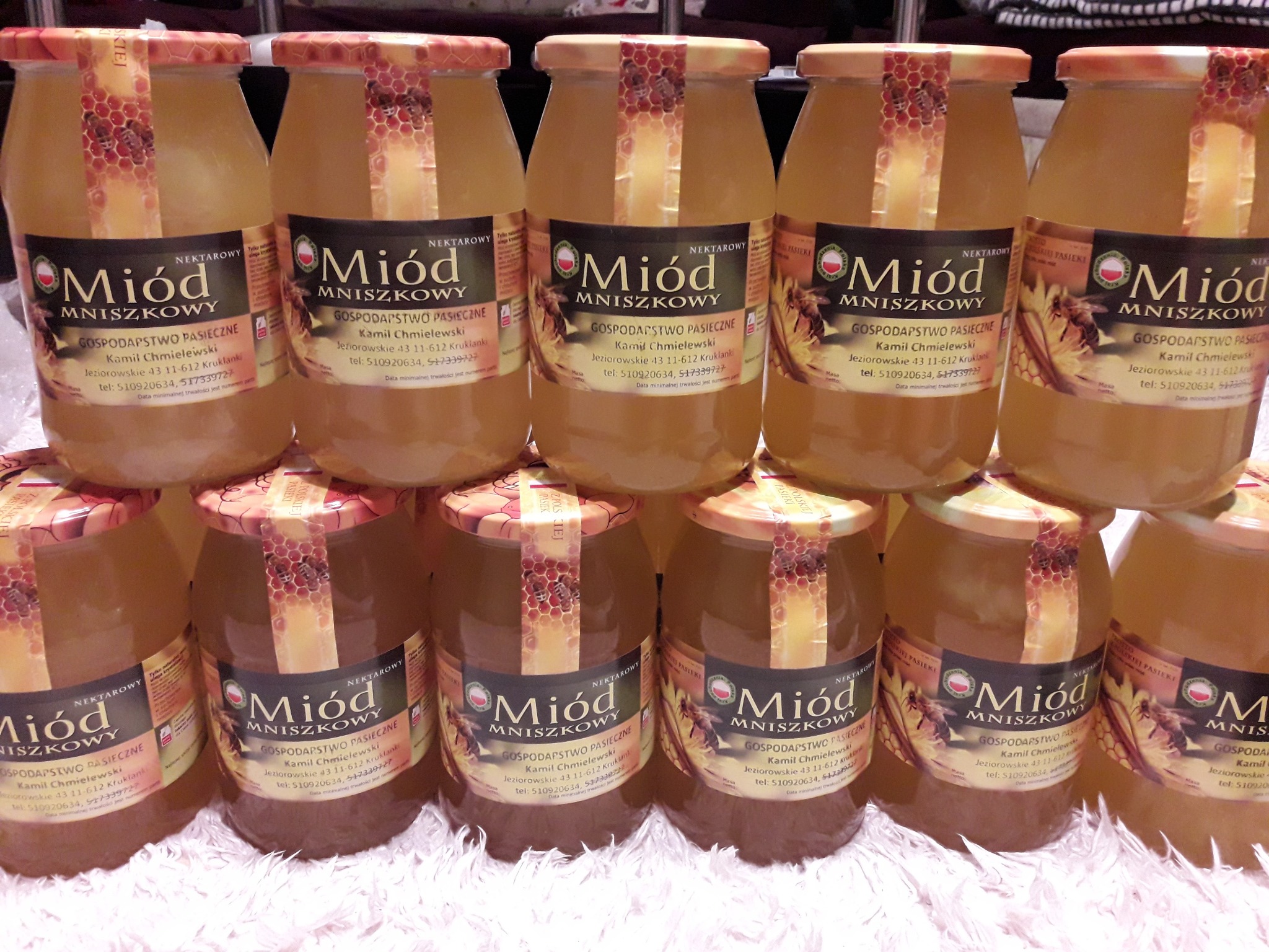 Ustawione piramidalnie słoiki z nektarowym mniszkowym, etykiety z nazwą gospodarstwa pasiecznego, widoczne numery telefonów i adres. Słoiki na jasnym, puszystym podłożu.