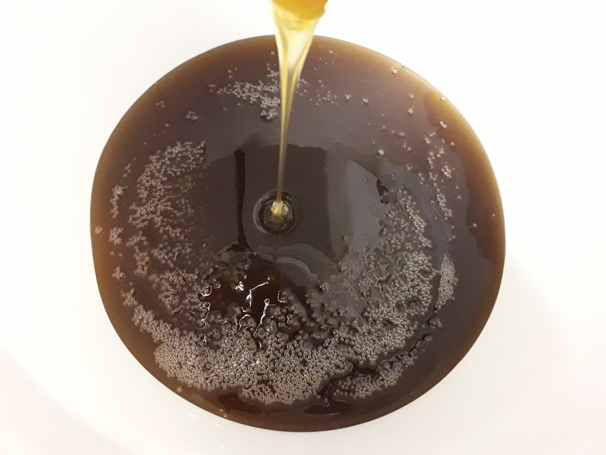Strumień bursztynowego płynu spływa do naczynia z ciemnym płynem, tworząc małe bąbelki na powierzchni.