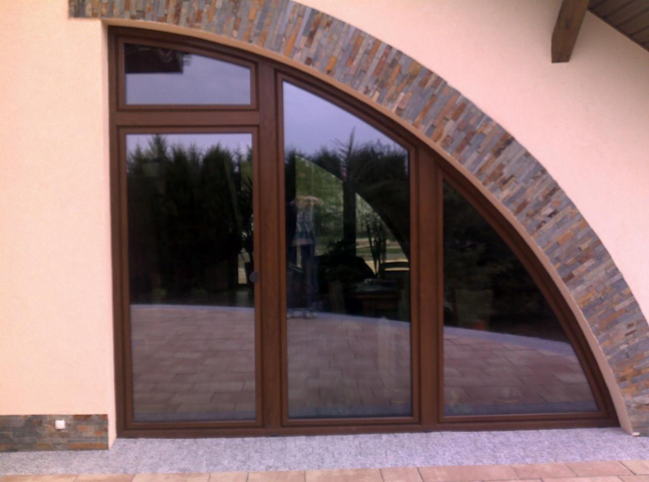 Brązowe okno PCV o nietypowym, łukowym kształcie, z kamienną obudową w odcieniach szarości i brązu, odbijające otoczenie ogrodu.