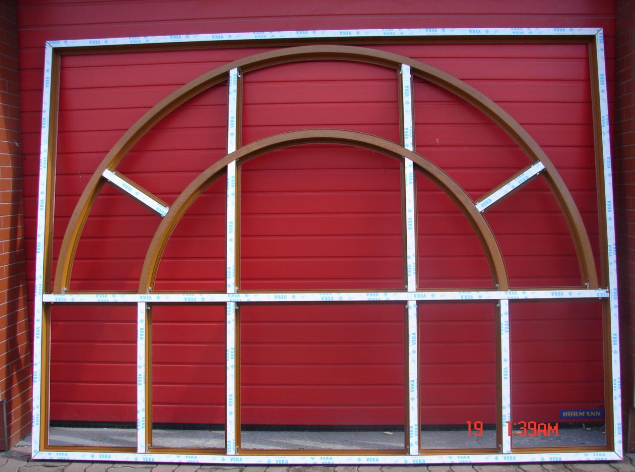 Szkielet okna z łukowym nadprożem, widoczne profile i elementy konstrukcyjne na tle czerwonej bramy garażowej.