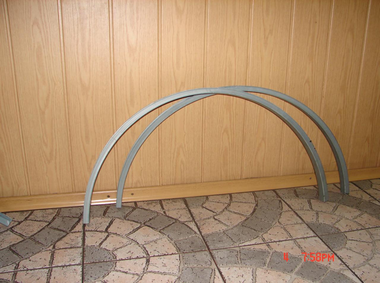 Dwa szare, metalowe łuki prawdopodobnie do konstrukcji okiennej, leżące na podłodze z płytek w odcieniach szarości i beżu, na tle boazerii w kolorze jasnego drewna.