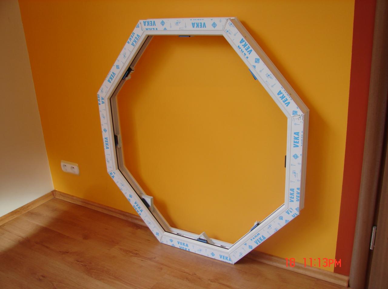 Ośmiokątna rama okienna z białego tworzywa z niebieskim nadrukiem VEKA, oparta o pomarańczową ścianę na drewnianej podłodze w trakcie montażu.