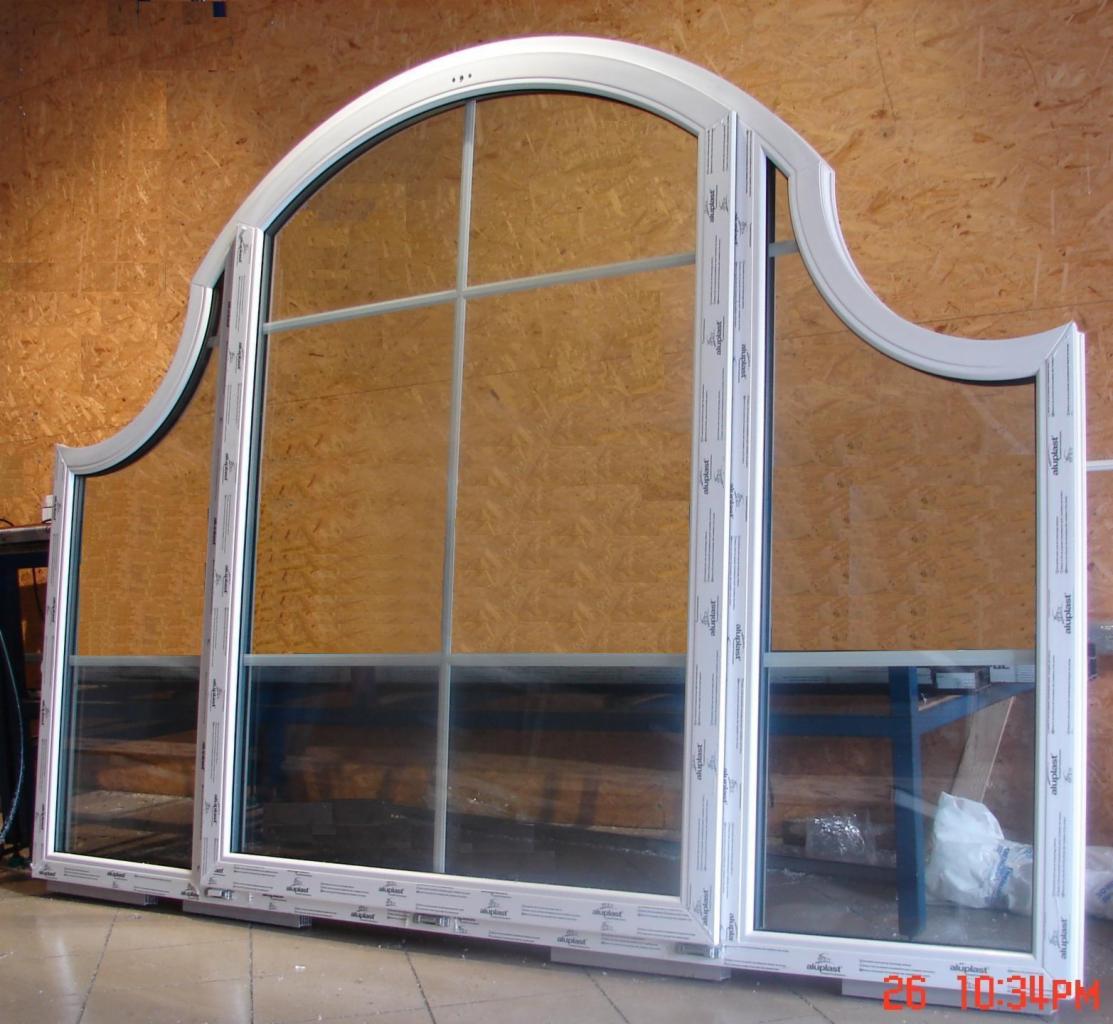 Trzyczęściowe okno z białymi ramami i łukowym górnym panelem, widoczne logo 'aluplast' na ramie, ustawione na podłodze, z drewnianą ścianą w tle.