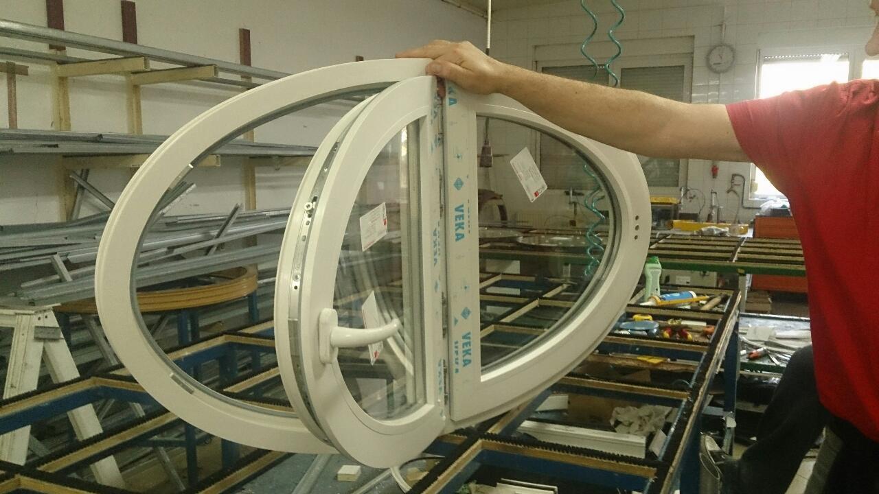 Owalne okno z uchylnym skrzydłem, w trakcie produkcji, widoczne oznaczenia producenta profili, na warsztatowym stole.
