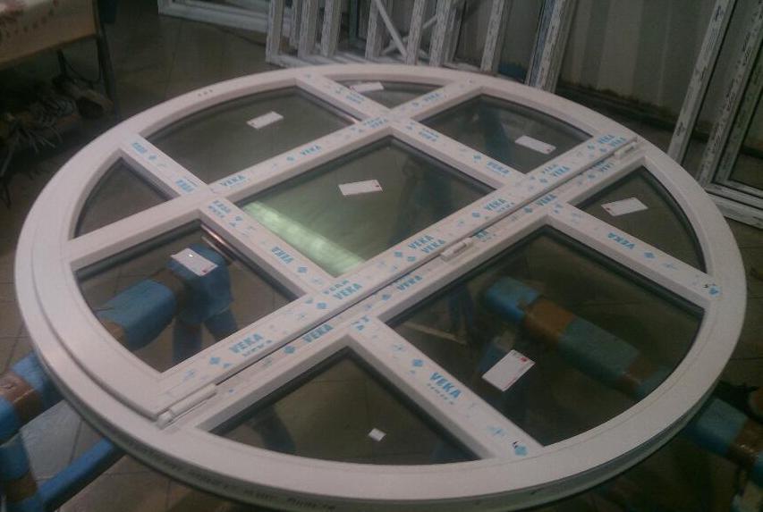 Okrągłe okno z białymi ramami z tworzywa sztucznego, podzielone na segmenty, z naklejkami producenta VEKA na profilach, leżące na zabezpieczonych rurach.