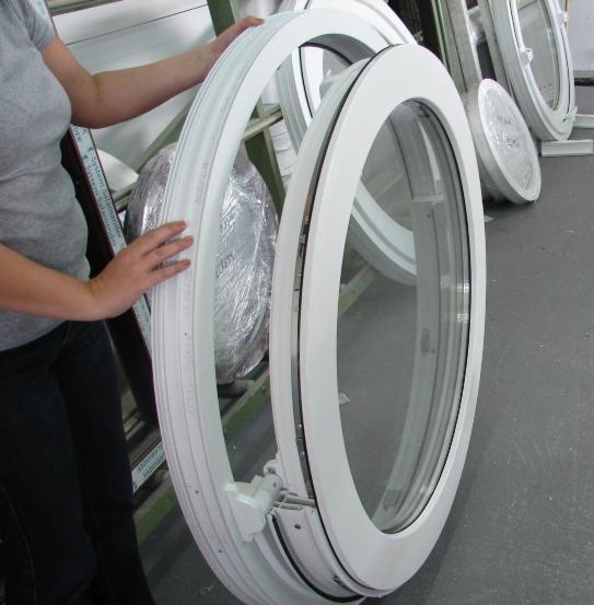 Owalne, białe okno uchylne, trzymane przez osobę, z widocznym mechanizmem otwierania, obok okna zapakowane w folię.