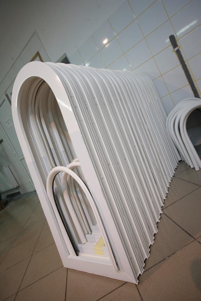 Stos białych, łukowatych ram okiennych PCV ułożonych w magazynie na kafelkowej podłodze, widoczne ślady transportu na dolnych ramach.