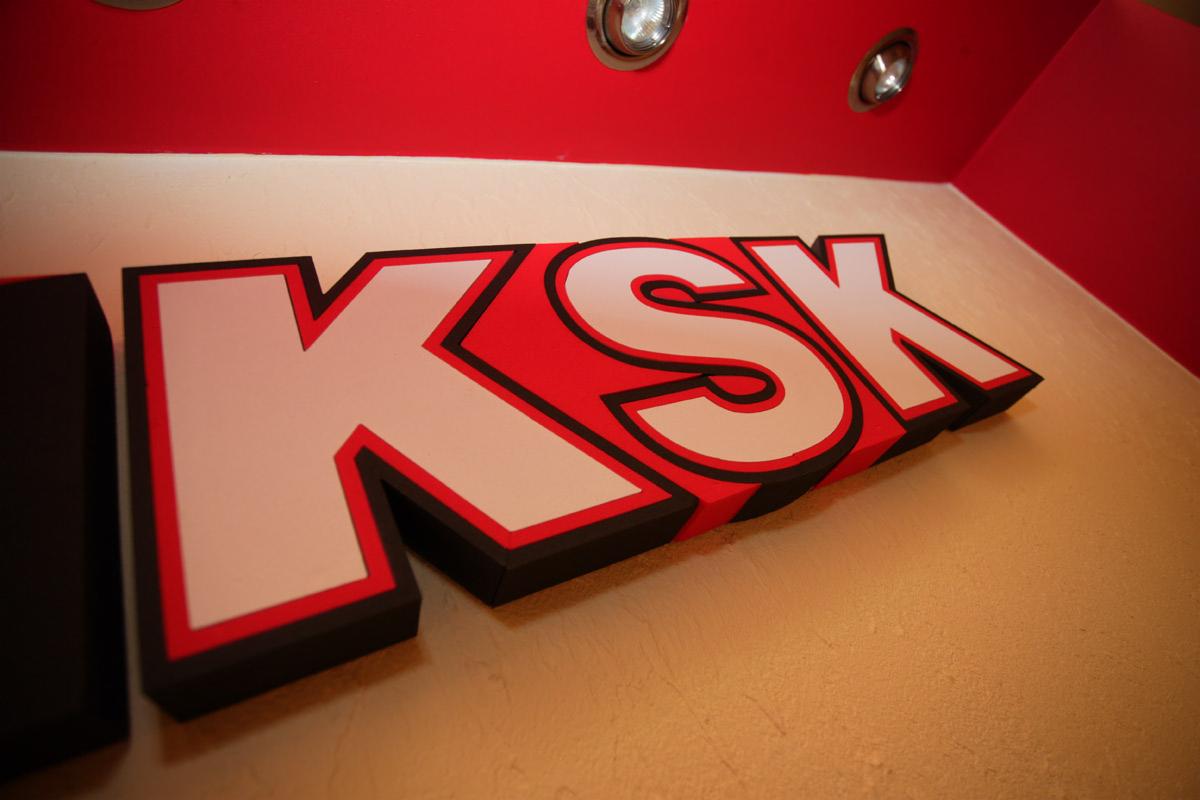 Trójwymiarowy, podświetlany znak 'KSK' w kolorach czerwonym, białym i czarnym, zamontowany na ścianie w rogu pomieszczenia z czerwonym sufitem i oświetleniem punktowym.