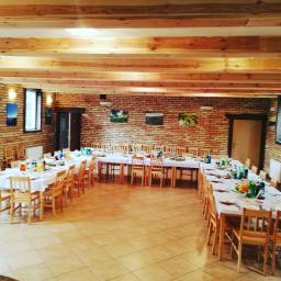 Tabaczarnia Sp. z o.o. - Sala bankietowa z długimi stołami nakrytymi białymi obrusami, drewnianymi krzesłami, przygotowana do przyjęcia gości, z ceglaną ścianą ozdobioną obrazami i drewnianym sufitem.