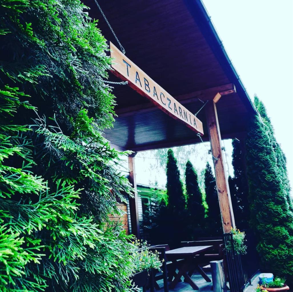 Zewnętrzny widok na zacieniony taras z drewnianym stołem i krzesłami, otoczony zielonymi tujami; tablica z napisem 'TABACZARNIA' zawieszona nad wejściem.
