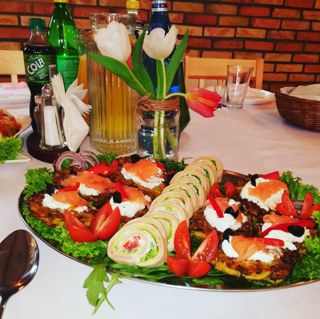 Dekoracyjnie podane dania na srebrnej tacy: roladki z szynki i warzyw oraz placki ziemniaczane z łososiem i kremowym serem, otoczone sałatą i pomidorami, w tle napoje i ceglana ściana.