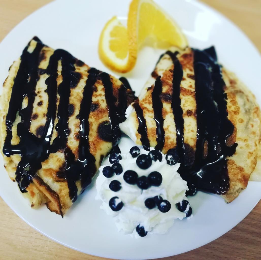 Dwa naleśniki polane ciemną polewą, podane z bitą śmietaną, borówkami i dwoma cząstkami pomarańczy na białym talerzu.