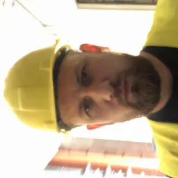 Autoportret brodatego mężczyzny w żółtym kasku i odblaskowej kamizelce na budowie, z widoczną instalacją elektryczną w tle. Ujęcie z dołu.