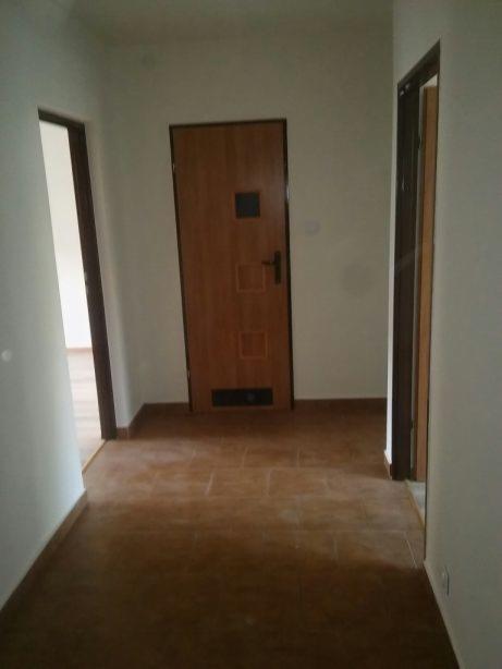 Korytarz z brązową podłogą z płytek, dwoje drzwi z ciemnymi futrynami i drewnianymi skrzydłami drzwiowymi z kwadratowymi zdobieniami, świeżo po remoncie, ściany pomalowane na biało.