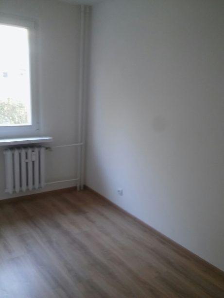 Pusty pokój po remoncie z panelami podłogowymi w odcieniach brązu, białymi ścianami, grzejnikiem pod oknem i widoczną instalacją grzewczą.