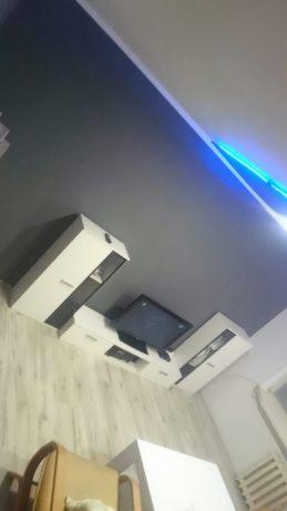 Nowoczesny salon z szarą ścianą i podświetleniem LED, białe meble modułowe z czarnymi wstawkami, widoczny fragment parkietu i krzesła.