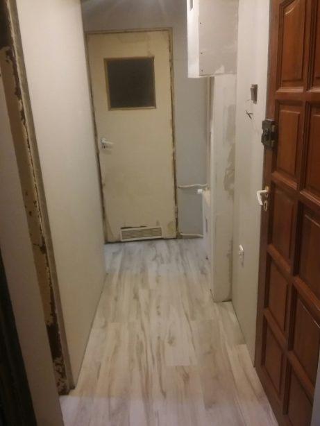 Korytarz w trakcie prac wykończeniowych: nowa podłoga imitująca drewno, stara, biała, metalowa futryna drzwi z okienkiem, ściany przygotowane do malowania, widoczne ślady po naprawach.