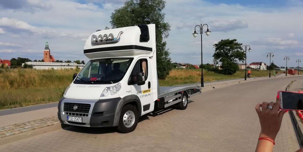 Biała laweta z nadbudówką i oświetleniem na dachu, zaparkowana na chodniku z widocznym numerem telefonu do kontaktu, w tle krajobraz miejski z kościołem i rowerzystą fotografowana telefonem.