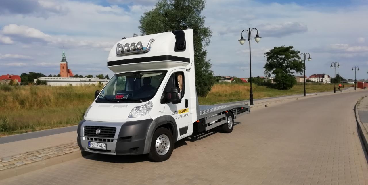 Biała laweta z kabiną sypialną i oświetleniem na dachu, zaparkowana na brukowanej drodze z widokiem na miasto w tle.