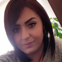 Portret kobiety z ciemnymi włosami i piercingiem w nosie, na tle widoczny fragment zielonej rośliny doniczkowej.