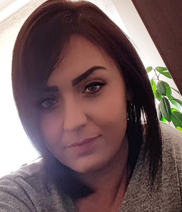 Portret kobiety z ciemnymi włosami i piercingiem w nosie, na tle widoczny fragment zielonej rośliny doniczkowej.